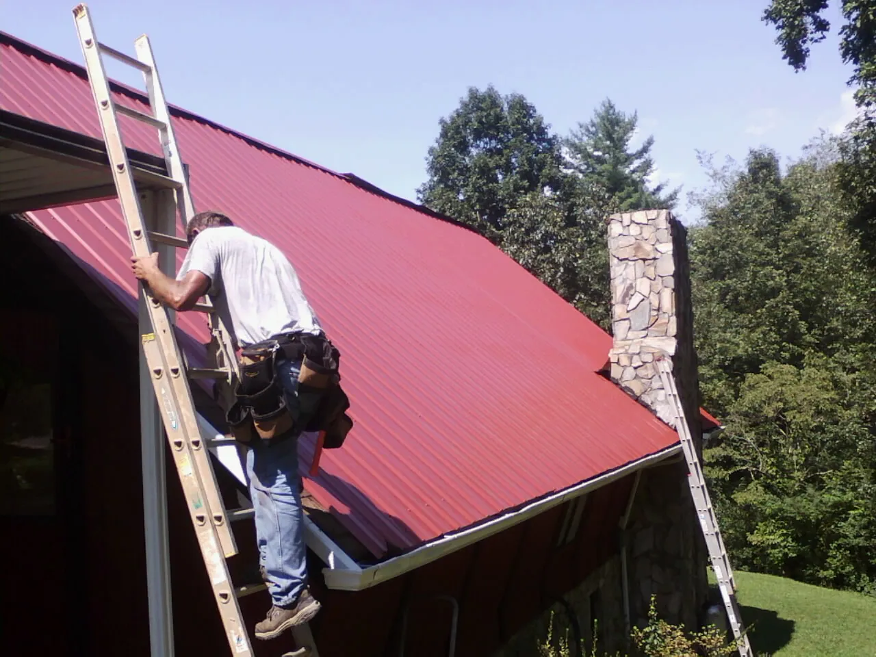 Roofer on a ladder beside a red metal roof and stone chimney — Soffit & Fascia Repair by Roofing Brothers in Haw River